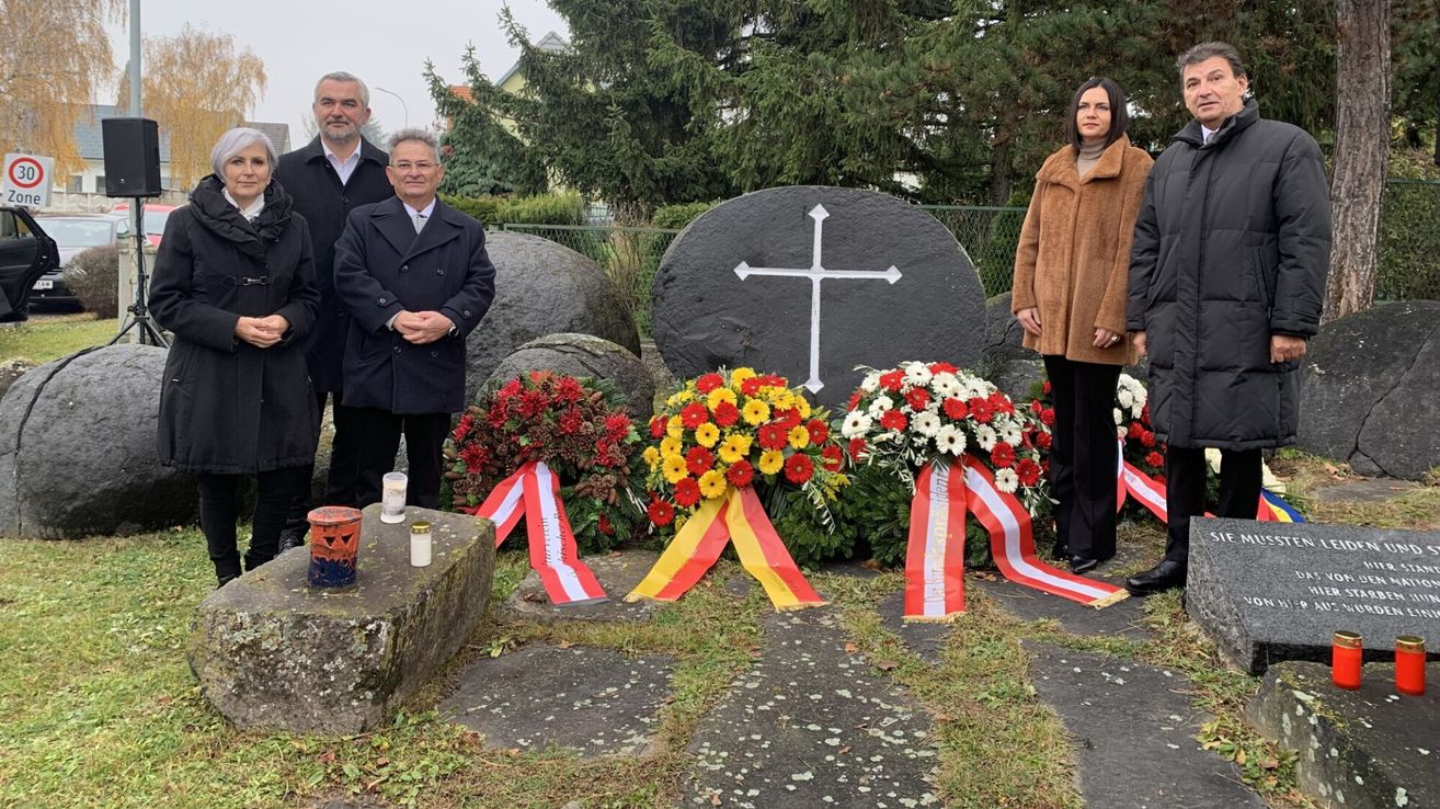 Three individuals stand before a memorial with a large cross and flowers. Two bouquets have red and white ribbons. A man and a woman are present.