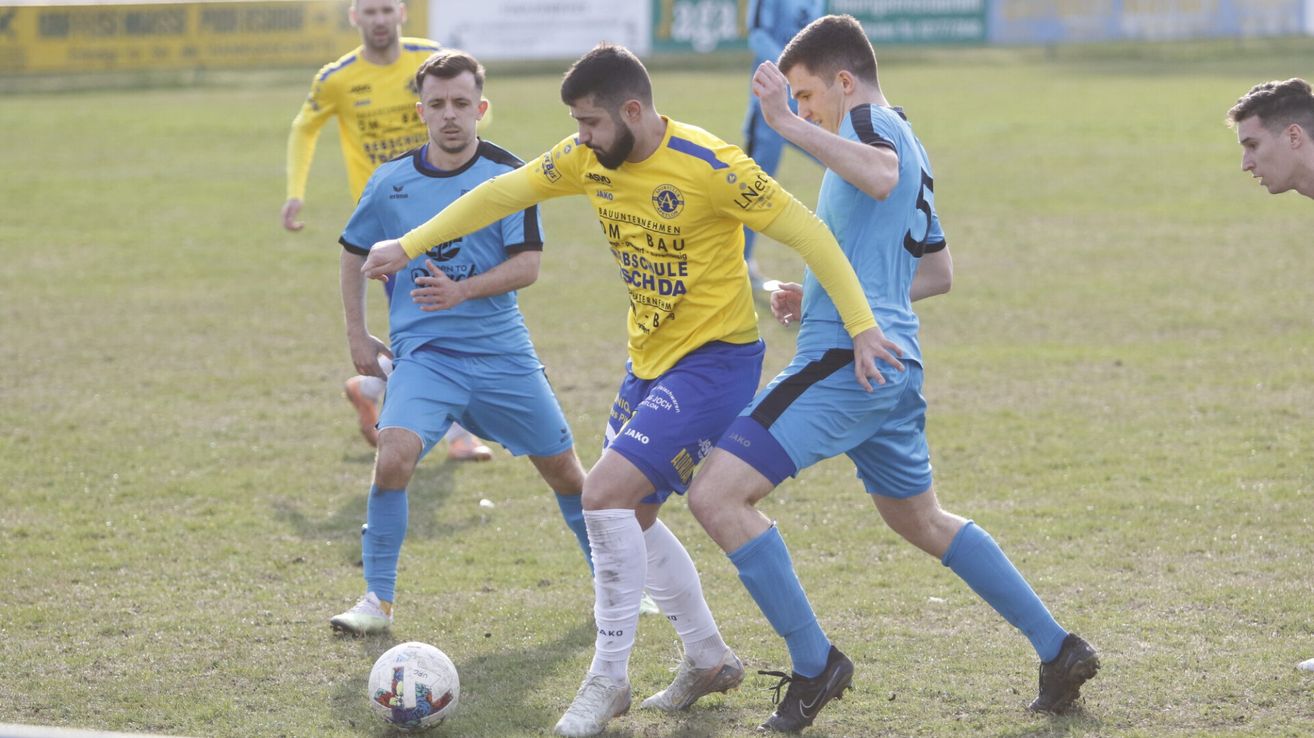Vier Fußballspieler sind auf einem Feld, zwei in Gelb und zwei in Blau. Der Spieler in Gelb versucht, den Ball zu schießen. Der Spieler in Blau versucht, ihn zu blockieren.