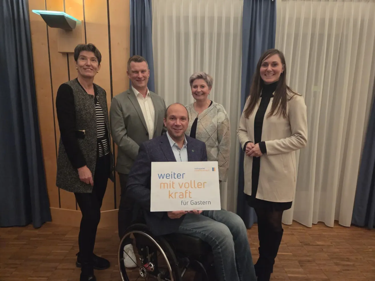 Five people, including a man in a wheelchair holding a sign, are standing in a room. The woman on the left wears a black and white striped top. The sign says, 'weiter mit voller kraft für Gasten.'