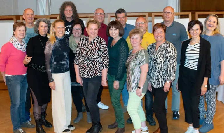 Eine Gruppe von Menschen steht auf einem Holzboden und lächelt für ein Foto. Einige tragen Brillen, und verschiedene Kleidungsstile sind sichtbar. Im Hintergrund ist ein Banner mit Text zu sehen.