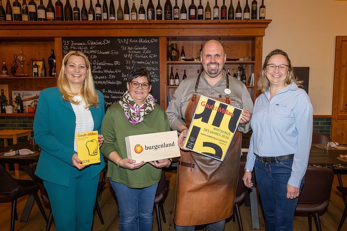 Vier Menschen posieren für ein Foto und halten Bücher und eine Schachtel in den Händen. Sie stehen vor einem Holzregal mit Weinflaschen. Der Mann in der Mitte trägt eine braune Schürze und hält ein gelbes Plakat. Die Frau links trägt einen grünen Blazer. Die Frau rechts trägt eine Brille und ein blaues Hemd.
