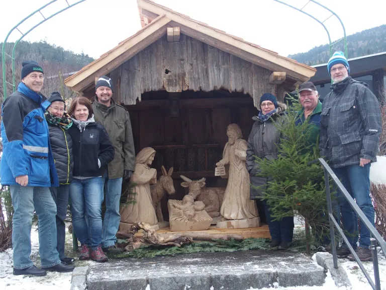 Eine Gruppe von sechs Personen steht vor einer hölzernen Krippe. Die Figuren umfassen Maria, Josef, das Jesuskind und Tiere wie Esel und Lamm.