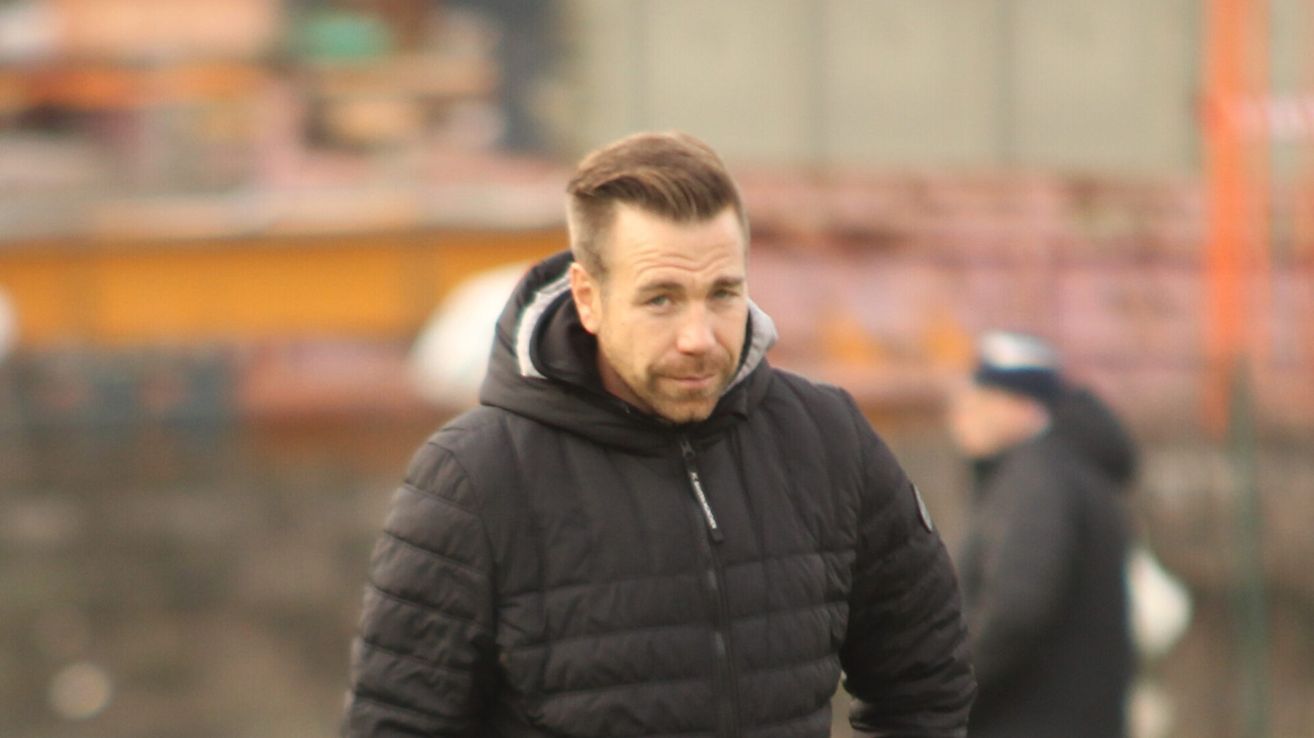 Ein Mann in einer schwarzen Jacke mit Kapuze und Reißverschluss steht an einem Ort im Freien, möglicherweise bei einer Sportveranstaltung.