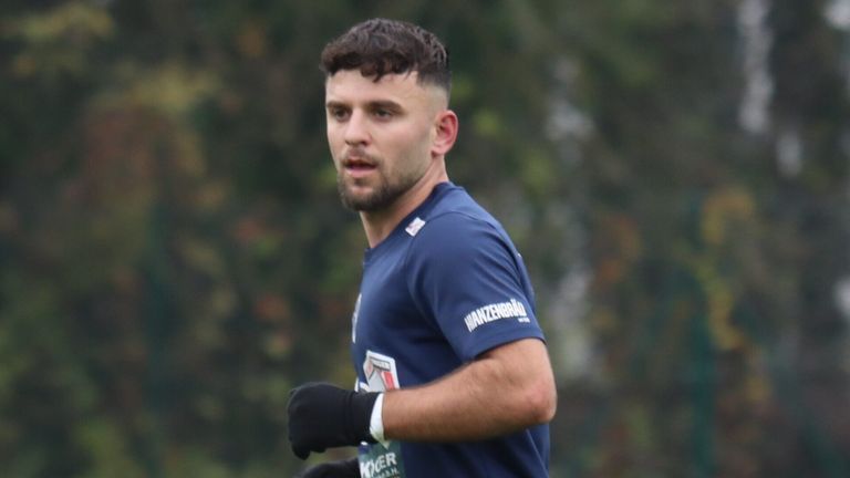 Ein Mann in einem blauen Trikot mit 'HANZENBRAU' darauf läuft im Freien, trägt schwarze Handschuhe und ein weißes Armband. Bäume sind im Hintergrund verschwommen.