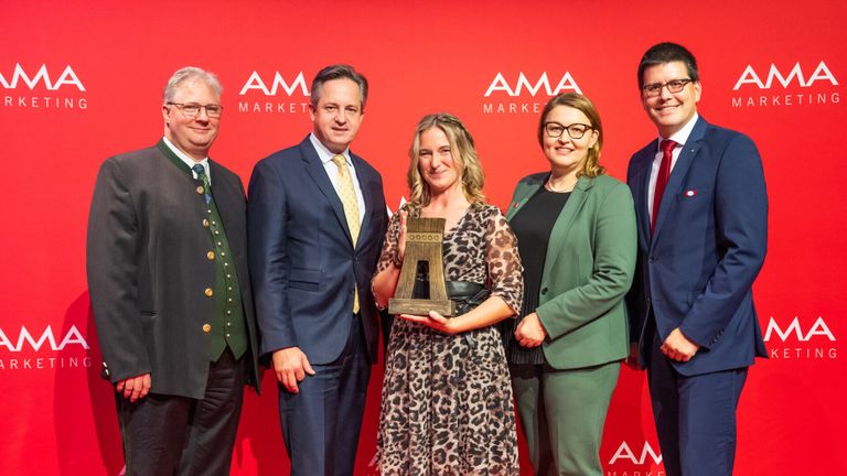 Five individuals stand in front of a red backdrop with logos. The central figure is a woman holding a wooden trophy. She is surrounded by two men and two women, all dressed formally.