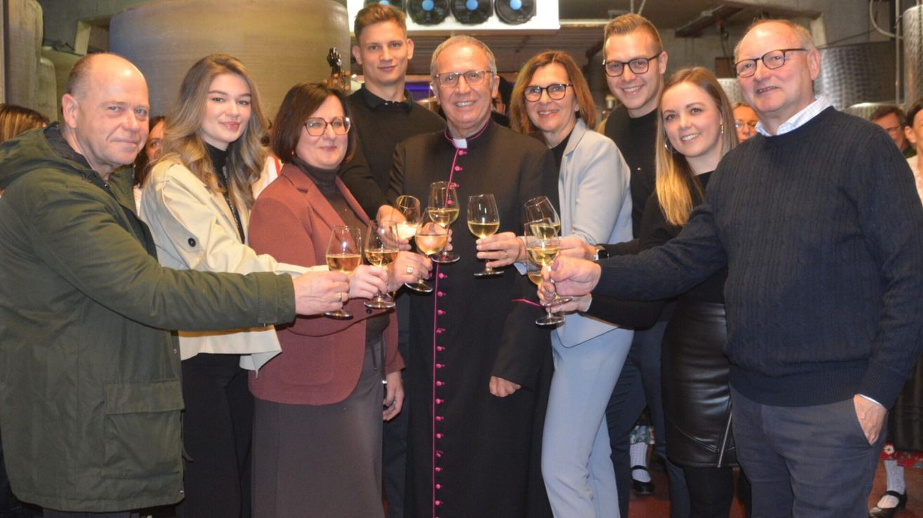 Eine Gruppe von Menschen, darunter ein Priester in schwarzen Gewändern, halten Weingläser und lächeln in die Kamera.