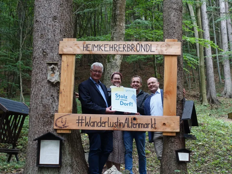 Vier Personen stehen in einem Wald und posieren vor einem Holzrahmen, auf dem 'Heimkehrerbruendl' steht. Eine Person hält ein Schild mit der Aufschrift 'Stolze auf unser Dorf!' und eine andere hält ein Hashtag-Schild.