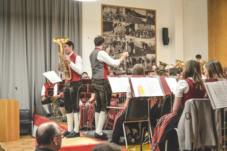 Eine Blaskapelle spielt auf der Bühne. Der Dirigent hält einen Taktstock und die Bandmitglieder spielen ihre Instrumente. Das Publikum sitzt vor ihnen.
