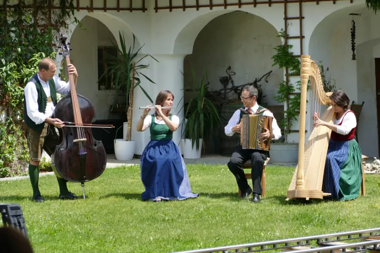 Eine Gruppe von Musikern spielt auf einem Rasen mit einem Bogen im Hintergrund. Eine Frau spielt Flöte, ein Mann spielt Cello und ein anderer Mann spielt Akkordeon.