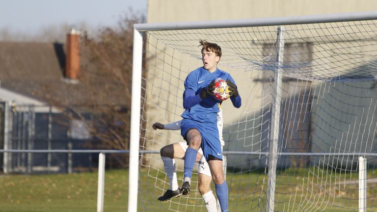 Ein Fußballspieler in blauem Trikot springt vor einem Tornetz und hält einen Fußball in der Hand.