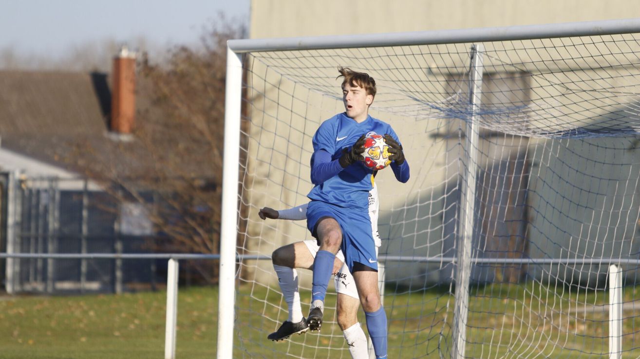 Ein Fußballspieler in blauem Trikot springt vor einem Tornetz und hält einen Fußball in der Hand.