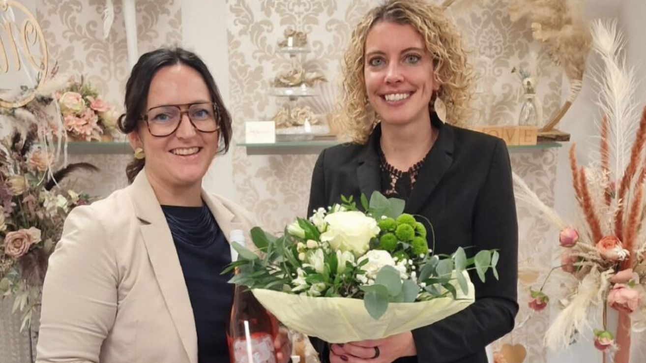 Zwei Frauen stehen vor einer dekorativen Wand. Die Frau auf der linken Seite hält eine Flasche Wein und die Frau auf der rechten Seite hält einen Blumenstrauß.