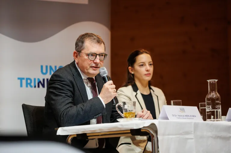 Ein älterer Mann spricht in ein Mikrofon auf einer Konferenz. Neben ihm sitzt eine Frau mit einem Glas Bier. Auf einem weißen Namensschild am Tisch steht 'Dr. Helmut Herlics'.