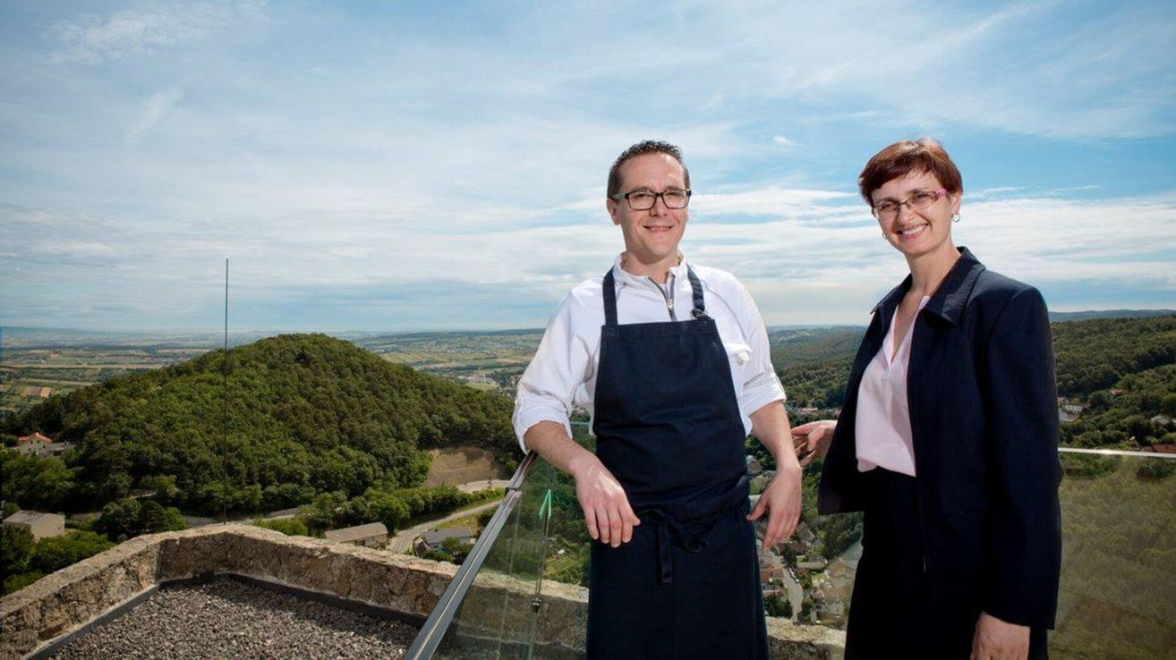 Ein Mann und eine Frau stehen lächelnd auf einem Balkon, der Mann in einem weißen Hemd und einer Schürze, die Frau mit Brille und einer rosa Bluse. Hinter ihnen ist ein Blick auf Hügel und eine Stadt zu sehen.