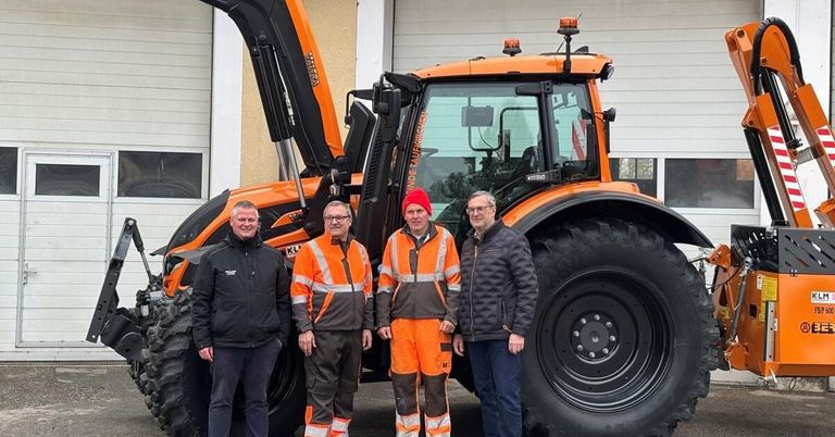 Vier Männer stehen vor einem orangefarbenen Traktor, zwei tragen reflektierende Westen, einer einen roten Hut und einer eine Brille. Hinter ihnen steht ein Gebäude mit weißer Wand und Glasfenstern.