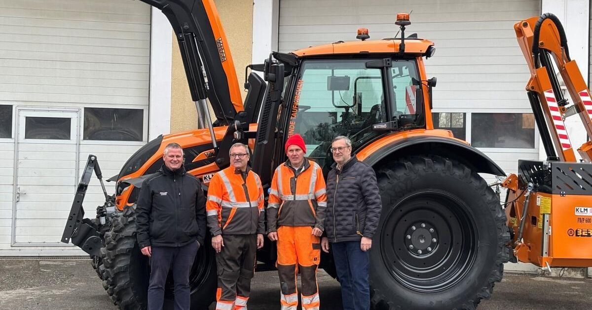 Vier Männer stehen vor einem orangefarbenen Traktor, zwei tragen reflektierende Westen, einer einen roten Hut und einer eine Brille. Hinter ihnen steht ein Gebäude mit weißer Wand und Glasfenstern.