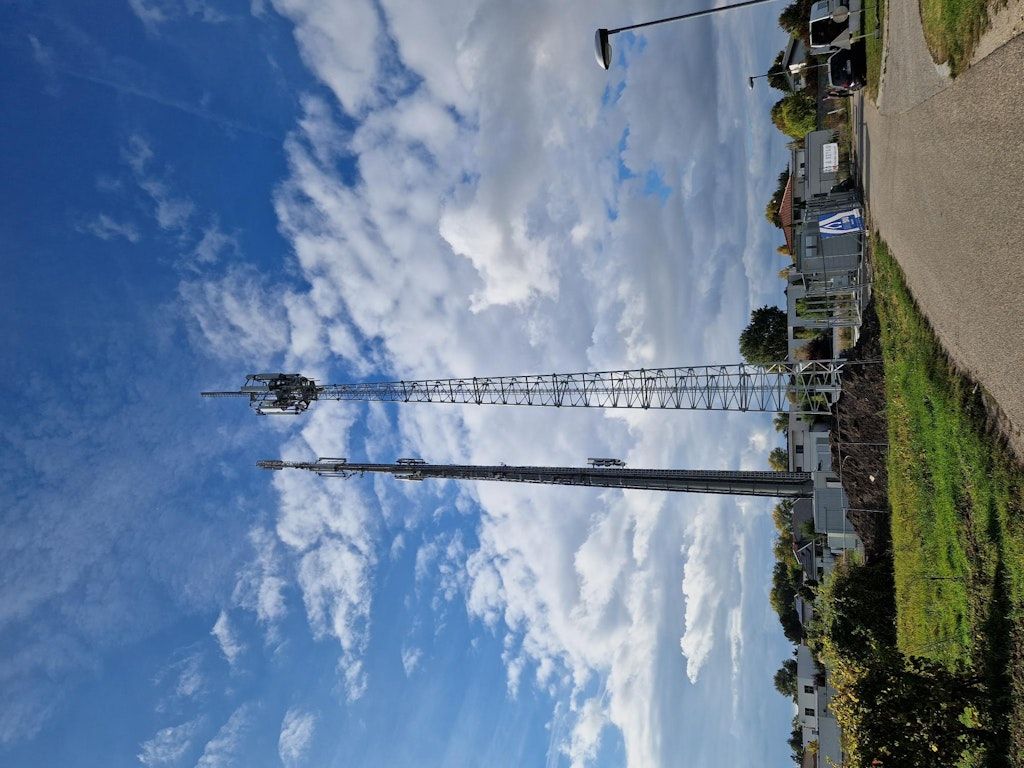 Ein Blick vom Boden zeigt zwei hohe Funktürme vor einem blauen Himmel mit weißen Wolken. Die Türme haben Metallstrukturen und sind von Grün umgeben.