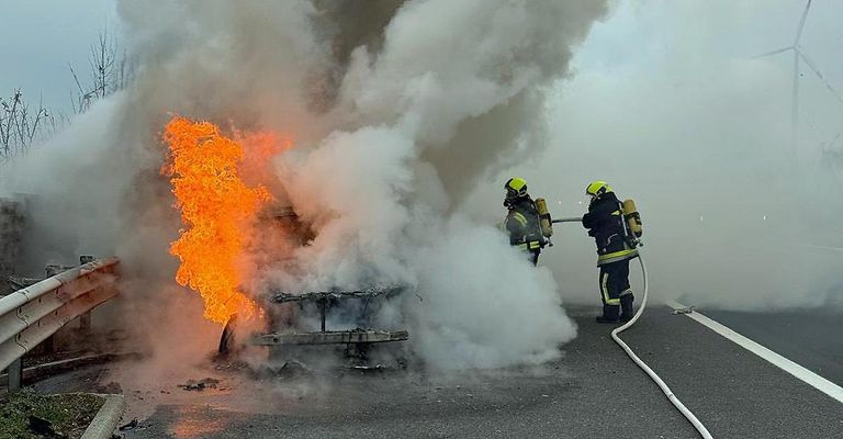 Zwei Feuerwehrleute löschen ein brennendes Fahrzeug auf einer Straße, wobei Feuer und Rauch sichtbar sind.