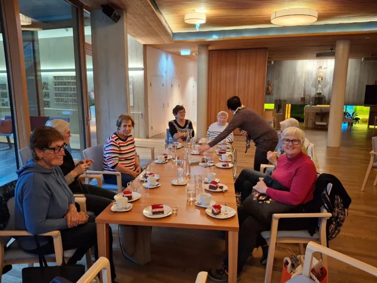 Eine Gruppe älterer Frauen sitzt um einen Tisch mit Dessertsplatten, eine Frau serviert sie. Der Raum hat Holzwände und eine Decke mit Lichtern. Es gibt Menschen, die auf Stühlen sitzen und um den Tisch herumstehen.