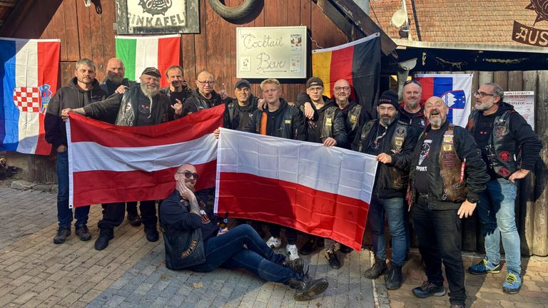 Eine Gruppe von Männern, einige in Lederjacken, posiert mit einer Flagge von Polen, Italien und Deutschland. Sie stehen und sitzen vor einem Gebäude mit einem Schild, das Cocktail Bar sagt.
