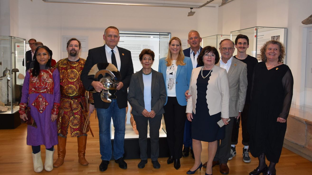 Eine Gruppe von Menschen steht in einem Museum und posiert für ein Foto. Ein Mann hält einen silbernen Helm, während ein anderer ein Kostüm trägt. Die anderen sind formell gekleidet. Dahinter befindet sich eine Vitrine mit einer Statue.
