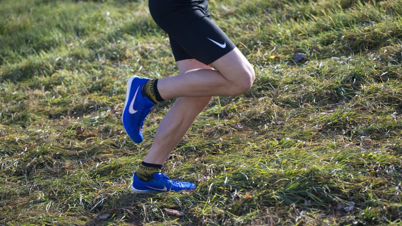 Eine Person läuft auf Gras und trägt blaue Nike-Schuhe und schwarze Nike-Shorts. Der Fokus liegt auf dem unteren Teil des Körpers.