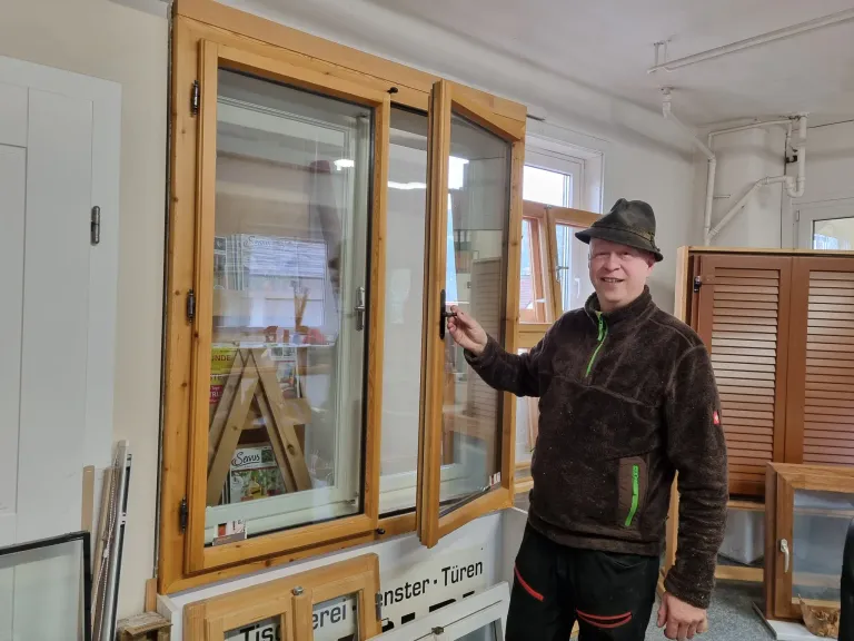 Ein Mann in einer braunen Jacke und Hut steht neben einem hölzernen Fenster und lächelt, mit Blick auf eine dreieckige Holzstruktur im Inneren.