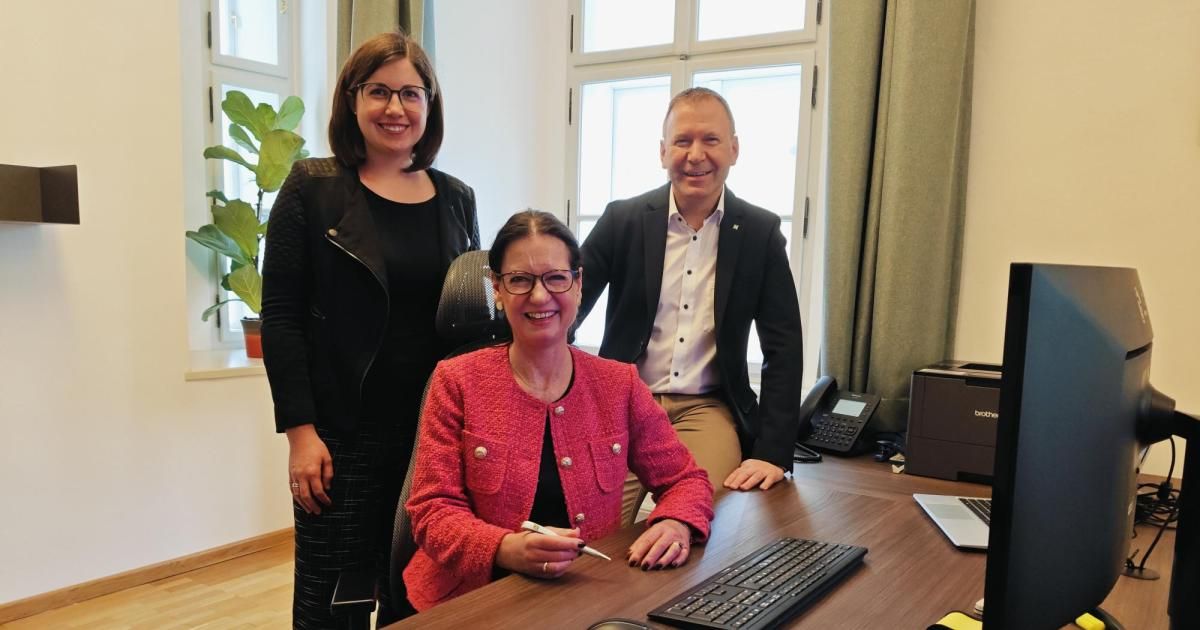 Drei Personen sind in einem Büro und lächeln für ein Foto. Eine Frau in einer rosa Blazer sitzt in einem Stuhl. Ein Mann steht hinter ihr und eine weitere Frau steht neben ihm. Der Schreibtisch hat einen Laptop, eine Tastatur und ein Telefon.