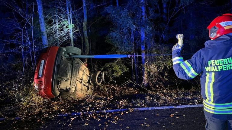Ein umgestürztes Auto am Straßenrand, bei Nacht, mit Rettungskräften, die ein Seil verwenden, um es aufzurichten. Das Auto ist rot, und seine Front ist zur Kamera gerichtet. Bäume und Büsche umgeben die Szene.
