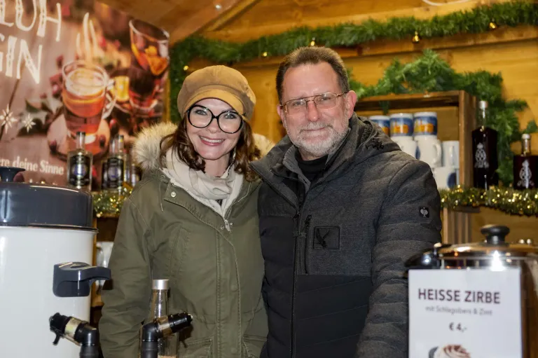 Ein Mann und eine Frau stehen nebeneinander und lächeln für die Kamera. Sie befinden sich in einem gemütlichen, festlichen Ambiente mit Holzwänden und Weihnachtsdekoration. Der Mann trägt eine Brille und eine dunkle Jacke, während die Frau eine Mütze und eine grüne Jacke trägt.