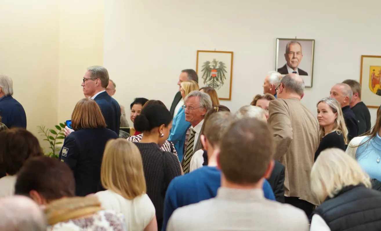 Eine Gruppe von Menschen, wahrscheinlich bei einer formellen Veranstaltung, steht und unterhält sich in einem Raum mit weißen Wänden und zwei gerahmten Bildern an der Wand.