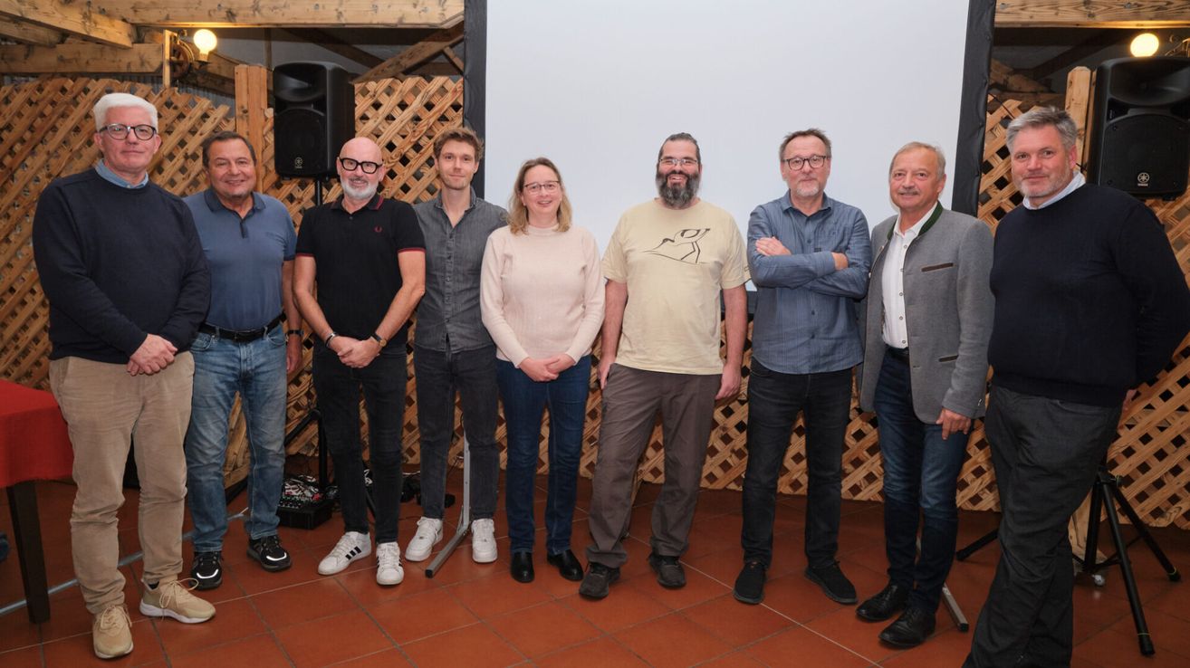 Eine Gruppe von sieben Personen, darunter Männer und Frauen, steht gemeinsam in einem Raum und lächelt für ein Foto. Der Raum hat ein hölzerne Gitter und eine weiße Wand im Hintergrund.