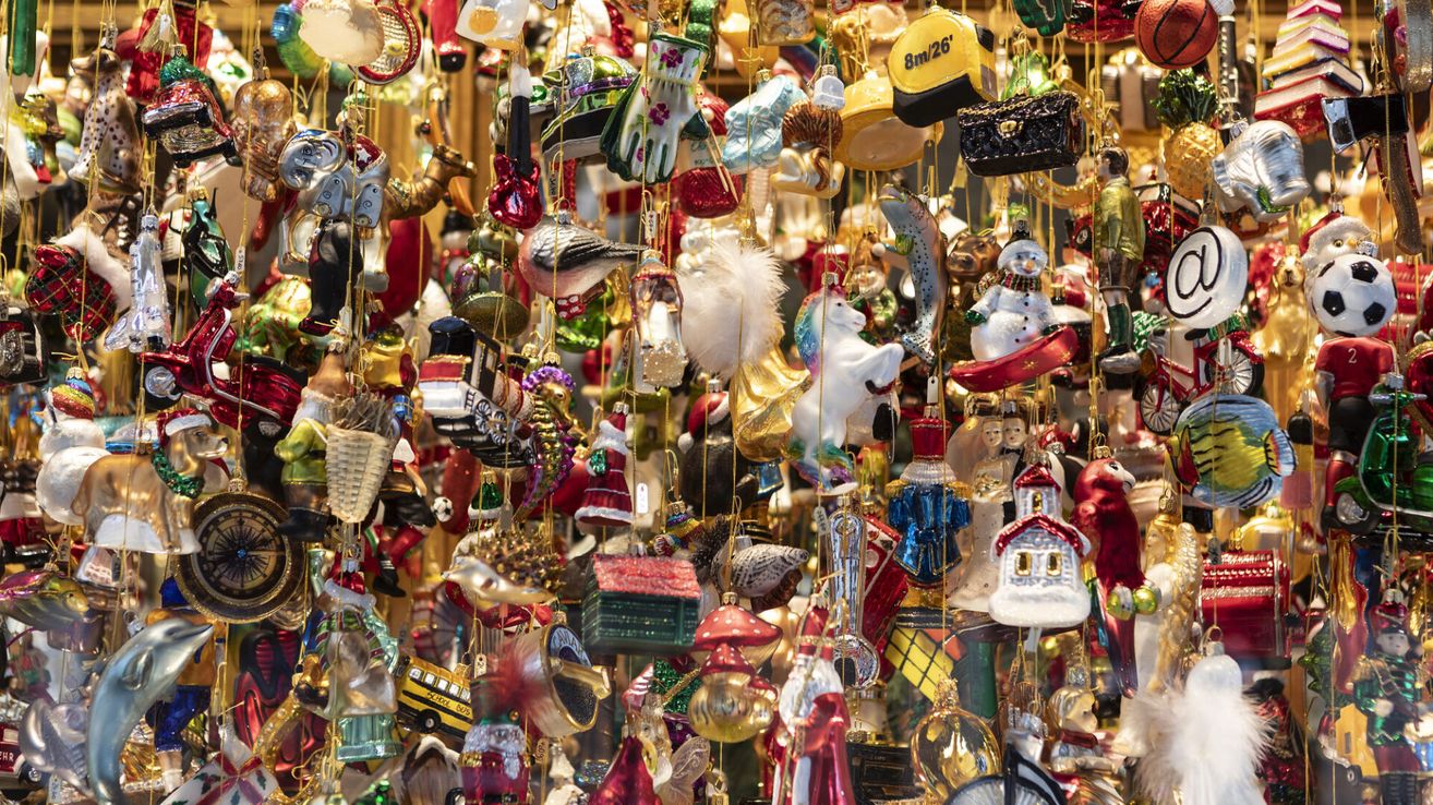 Eine farbenfrohe Ausstellung von Weihnachtsschmuck in verschiedenen Farben und Designs hängt an Schnüren. Bemerkenswerte Ornamente sind ein Schneemann, ein Einhorn und eine Werkzeugkiste. Die Ornamente spiegeln einen festlichen Weihnachtsgeist wider.