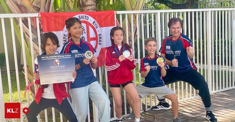 Fünf Kinder posieren mit Medaillen und Urkunden vor einem weißen Zaun. Hinter ihnen befindet sich ein Banner mit der Aufschrift Shito Ryu GNAS.