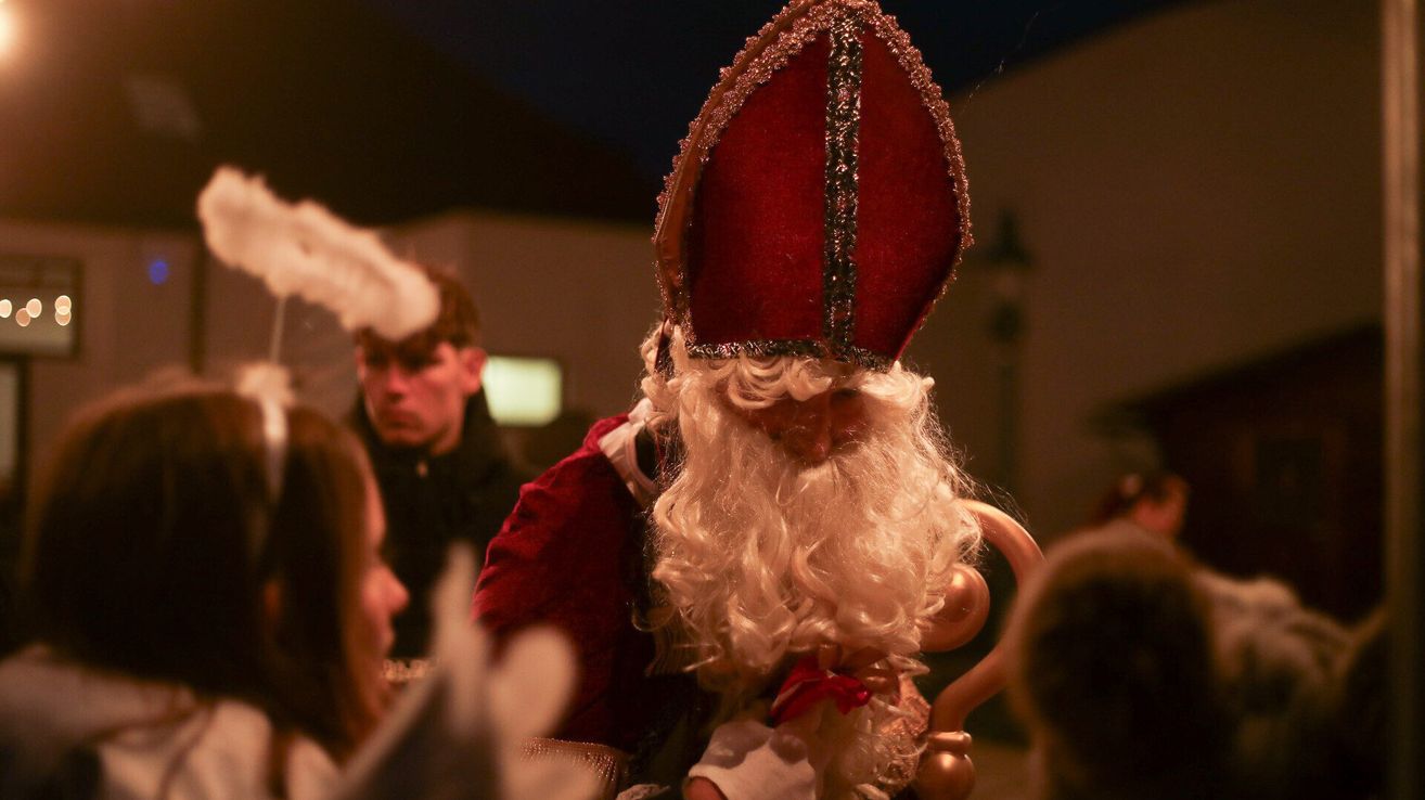 Eine Nahaufnahme einer Person, die als Weihnachtsmann gekleidet ist, einen goldenen Stab hält und einen roten Bischofshut trägt. Im Hintergrund sind verschwommen andere Personen zu sehen, eine mit weißen Federn.