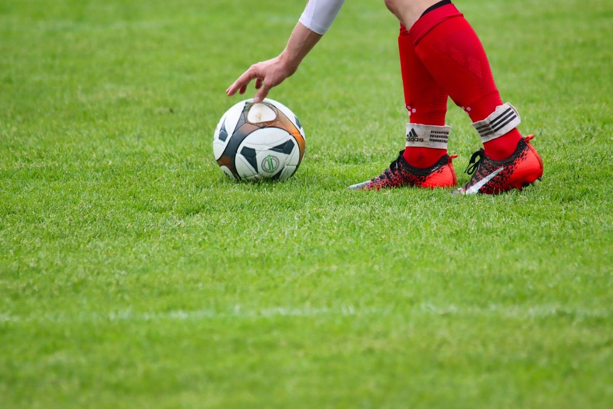 Ein Fußballspieler in roten Shorts und Socken berührt einen Fußball auf einem grünen Rasen.