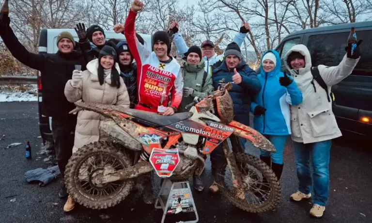 Eine Gruppe von Menschen steht neben einem schmutzigen Motocross-Motorrad, trägt Winterkleidung und einige haben Mützen auf. Der Mann in der Mitte hebt seine Hände, und das Motorrad hat Sponsoren und Text darauf.