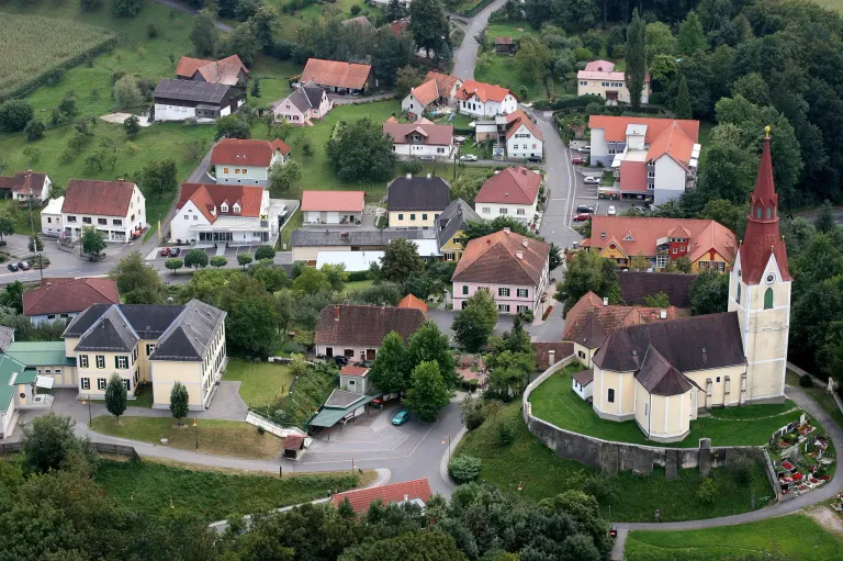 Luftaufnahme eines Dorfes mit zahlreichen Häusern in verschiedenen Farben, umgeben von grünen Feldern und Bäumen, mit einer zentralen Straße und einer Kirche mit einem Turm.
