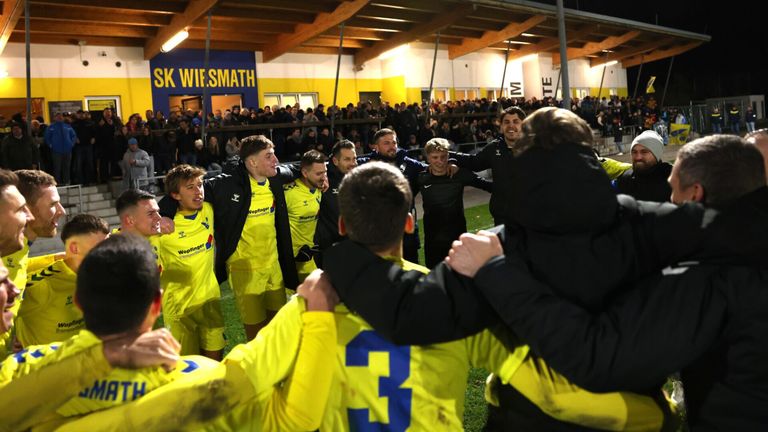 Fußballspieler in gelben Trikots stehen zusammen im Kreis auf dem Feld, während eine Menge im Hintergrund zuschaut.