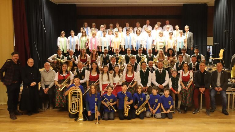 Eine große Gruppe von Musikern und Sängern in festlicher Kleidung, einschließlich traditioneller Kleidung, posiert für ein Foto auf der Bühne.