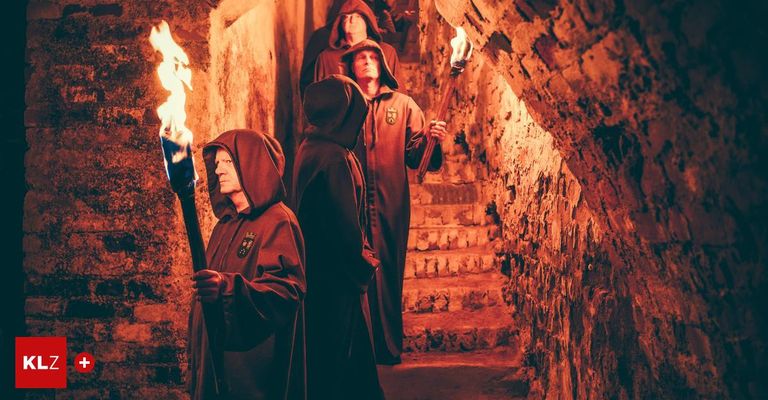 Vier Männer in Roben mit Fackeln stehen auf Steinstufen, zwei von ihnen blicken nach vorne. Die Fackeln sind entzündet, und die Atmosphäre ist düster.