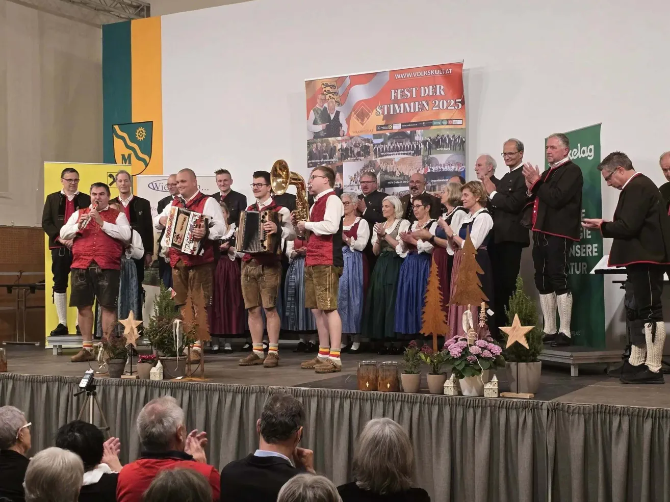 Ein Chorauftritt auf einer Bühne mit einem Banner, das Fest der Stimmen 2025 steht. Verschiedene Menschen klatschen und jubeln.