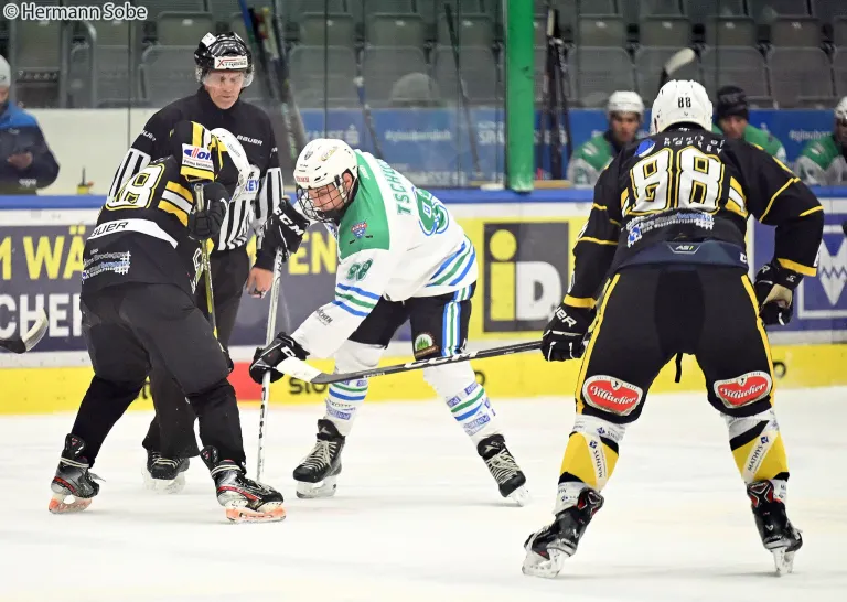 Eishockeyspieler in Uniform spielen auf einer Eisbahn. Ein Spieler hält einen Schläger, während ein anderer zuschaut. Die Eisbahn hat leere Sitze im Hintergrund.