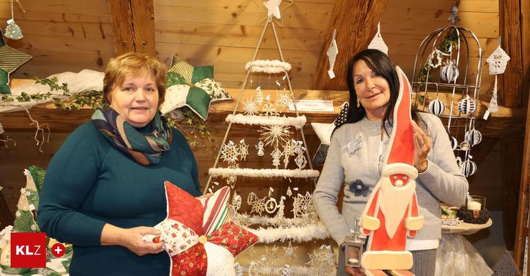 Zwei Frauen stehen vor einem hölzernen Hintergrund mit Weihnachtsdekoration. Eine Frau hält eine Weihnachtsmannfigur, und die andere hält ein Weihnachtskissen. Der Weihnachtsbaum hat ein weißes und grünes Farbschema.