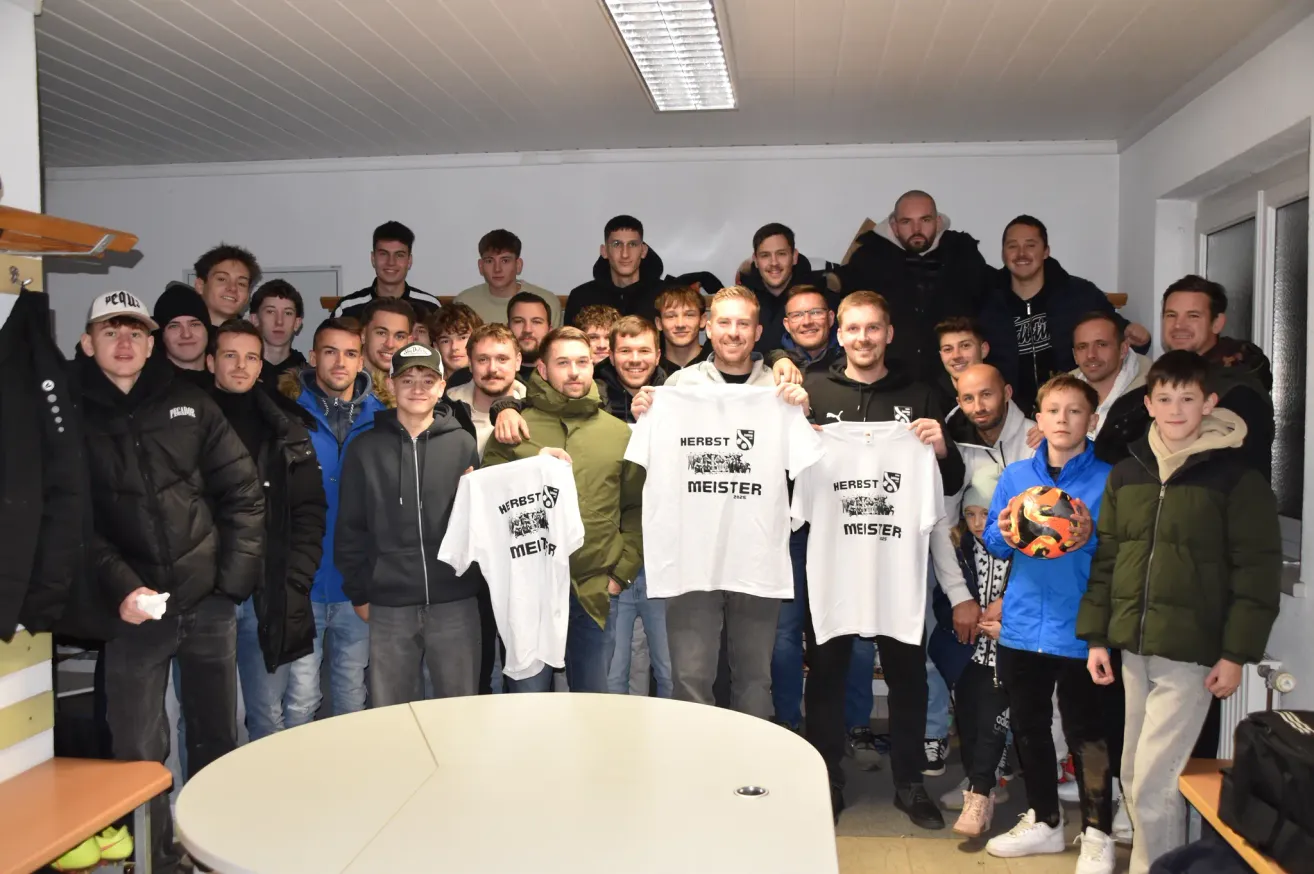 Eine Gruppe junger Männer und älterer Männer posiert für ein Foto, einige halten weiße T-Shirts mit der Aufschrift 'HERBST MEISTER' hoch. Der Raum ist hell mit weißen Wänden und einer Decke.