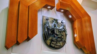 A metal plaque with a heraldic eagle design is mounted on a white wall. It features a cross on its chest and is enclosed by a wooden frame with embedded lights.
