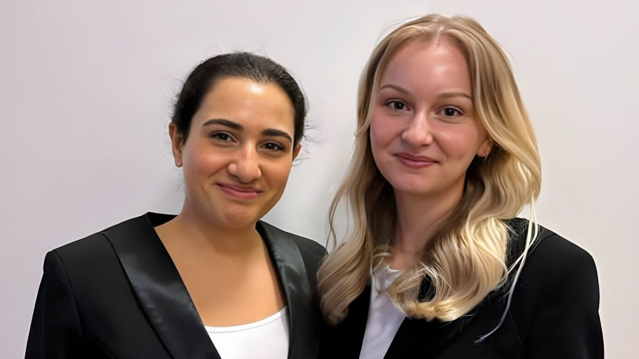 Two women standing next to each other, both smiling, against a plain background. The woman on the left has short dark hair and is wearing a black jacket. The woman on the right has long blonde hair and is wearing a black blazer.