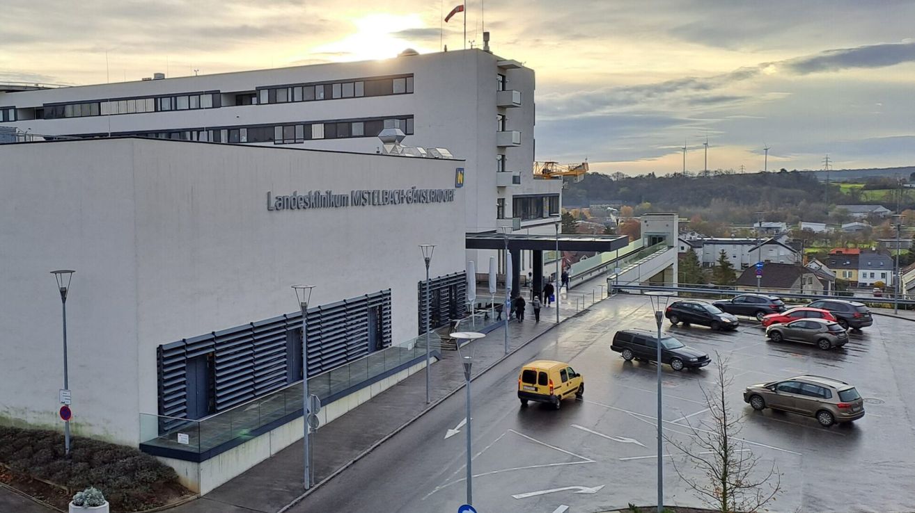 A hospital named Landesklinikum Mistelbach-Gänserndorf is visible, with cars parked outside. A yellow car is driving, and people are walking. The sky is cloudy.