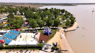 An aerial view of a beach area with a volleyball court, tents, and people walking on the sand. Trees and buildings are visible in the distance.