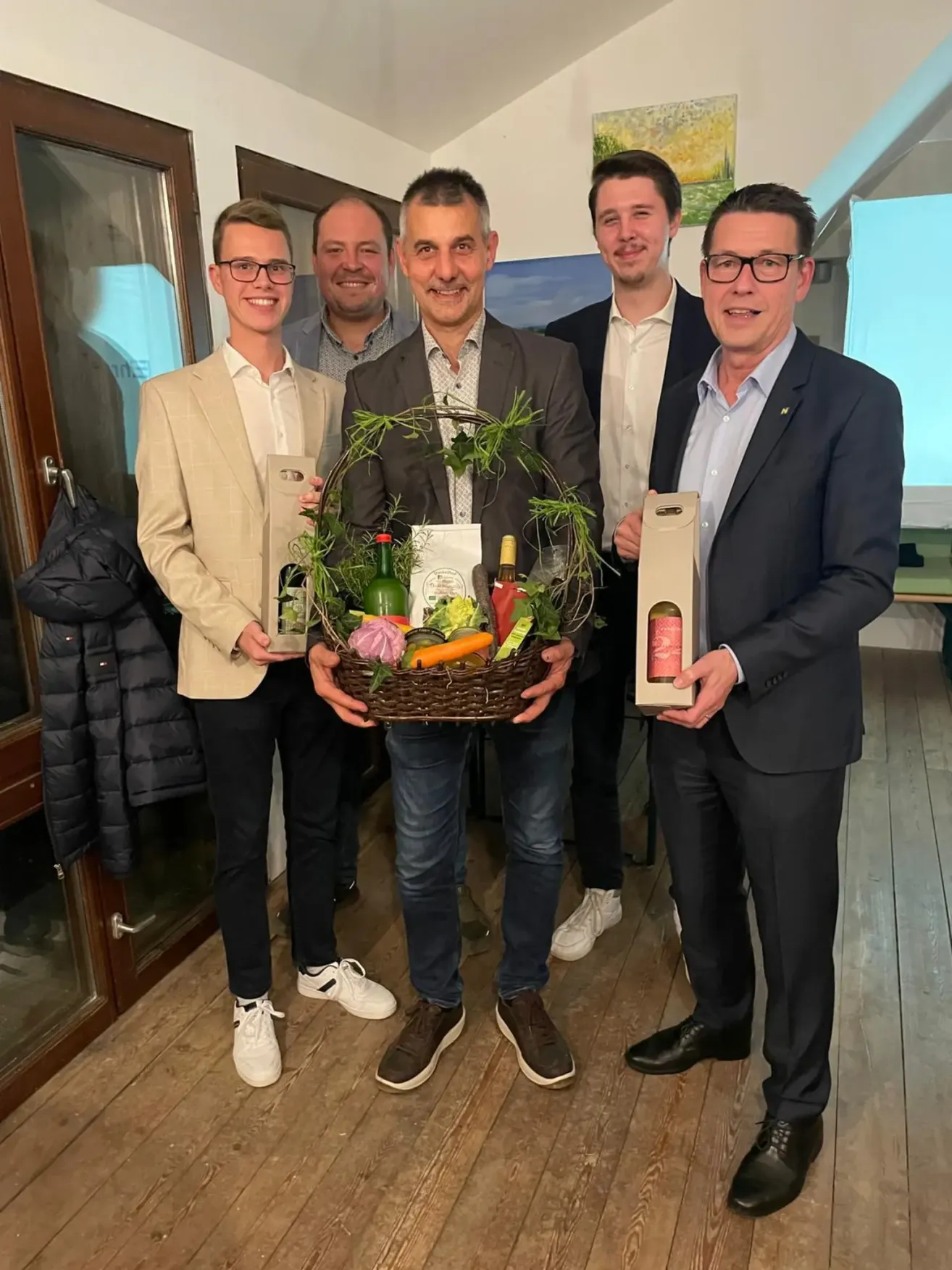 Five men stand in a room holding a basket of vegetables and bottles. The man in the center smiles, and the others look on. Behind them, a door is slightly open.
